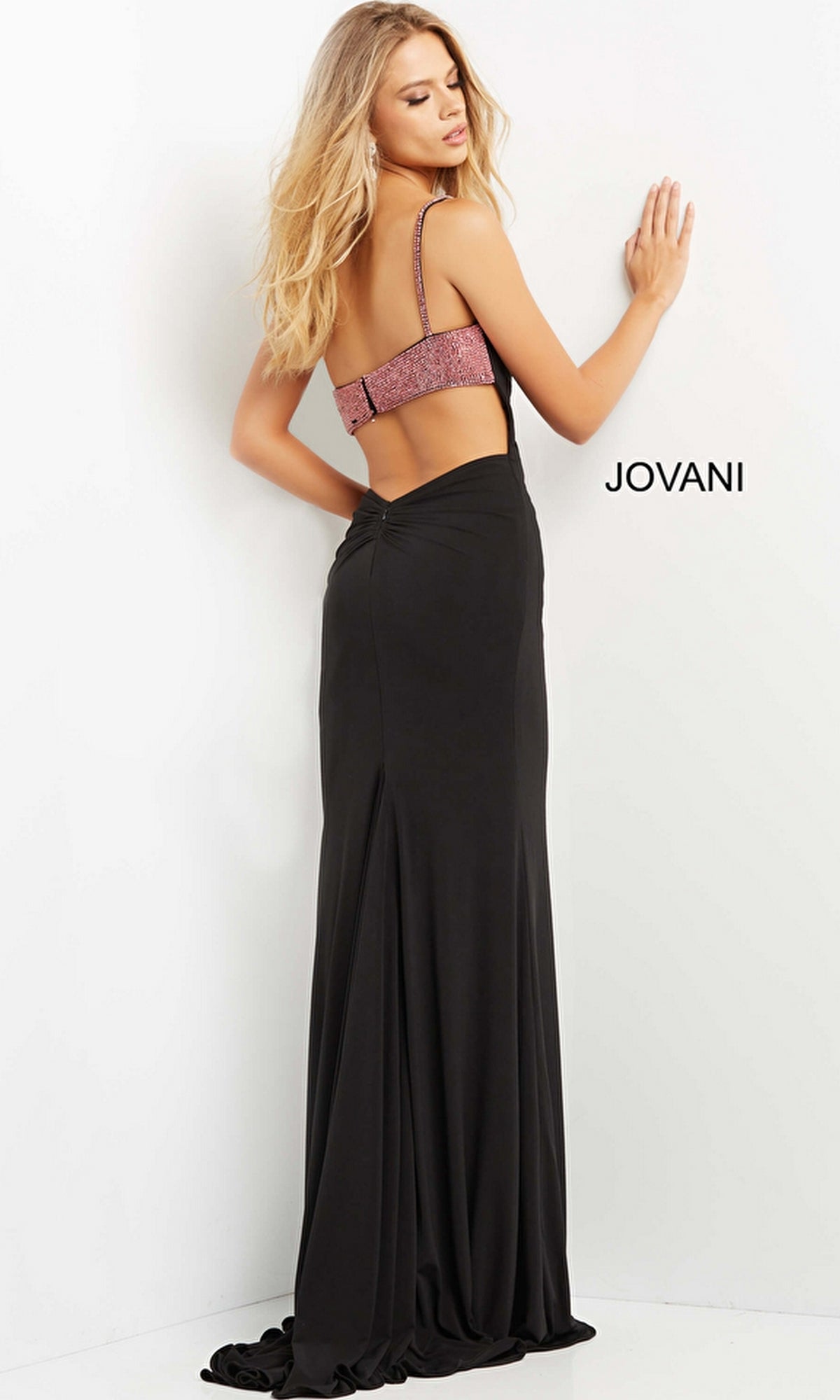 A woman poses against a white wall in the Jovani One-Shoulder Two-Tone Long Prom Dress 09021—a stylish black gown with an open back and sparkly pink strap—exuding modern elegance. The brand
